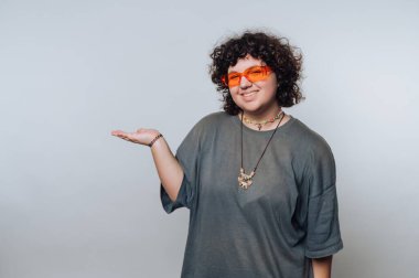 A young adult with curly hair and colorful accessories stands confidently, smiling and extending one hand outward. The neutral background highlights their cheerful expression.