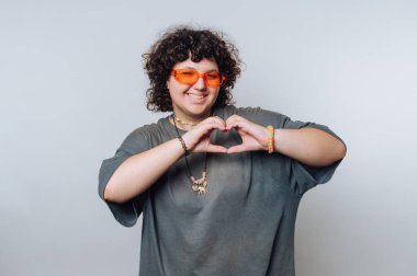 A cheerful individual with curly hair shows love by forming a heart shape with their hands. They wear bright orange glasses and have a casual, relaxed appearance.