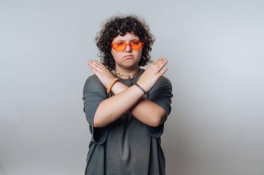 A young individual with curly hair wears stylish orange sunglasses and crosses their arms in a confident stance. The background is a simple light gray, creating a focused look.