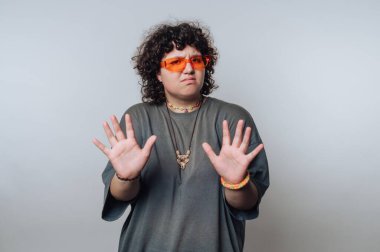 A person with curly hair stands against a plain background wearing orange sunglasses and a grey shirt. Their hands are raised in a stopping gesture, expressing a playful mood.