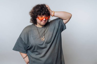 A person with curly hair stands against a plain light background, wearing oversized orange glasses. They have a playful pose and are dressed in a loose gray shirt. Colorful accessories add flair.