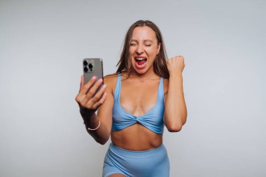 A joyful woman is posing with a smartphone, exuding excitement with a big smile. She is wearing a light blue fitness outfit, celebrating a positive moment in a simple indoor setting.