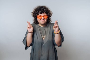 A person with curly hair is standing against a plain background, wearing bright orange sunglasses and a comfortable outfit. They smile and cross their fingers, expressing optimism.