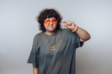 A young individual stands confidently, showcasing orange sunglasses while making a playful gesture with their fingers. The backdrop is simple, allowing their personality to shine through.