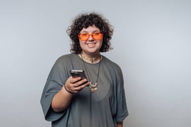 A cheerful individual with curly hair is standing in front of a plain background. They are wearing orange sunglasses and holding a smartphone, enjoying a moment of leisure.