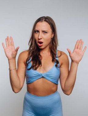 A woman in a blue crop top and matching leggings raises her hands in the air, seemingly surprised or excited. The background is light and simple, focusing on her expression.