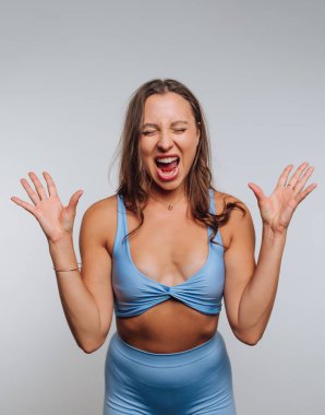 A woman is joyfully posing in a light blue workout outfit, with her hands raised and a big smile on her face. The bright studio setting emphasizes her vibrant energy.