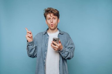 A young man in a denim jacket holds a smartphone with a surprised expression. He stands in front of a blue wall, indicating an unexpected discovery or realization.