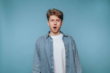 A young man stands in front of a blue background, wearing a denim shirt and a white t-shirt. He has a shocked expression, mouth open in surprise, as if taken aback by unexpected news.
