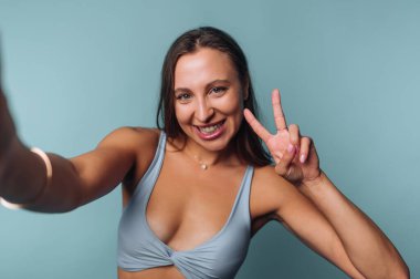 A young woman smiles and gives a peace sign while taking a selfie. She wears a light blue crop top, showing a joyful demeanor against a soft blue backdrop.