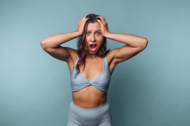 A woman stands in front of a light blue background with her hands on her head, showing a shocked expression. She wears a matching blue top and bottoms, emphasizing her surprised look.
