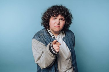 A person with curly hair is pointing directly at the viewer, wearing a casual jacket over a white shirt. The background is a solid light blue, enhancing the focused expression and gesture.