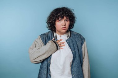 A young person with curly hair and a denim jacket looks surprised. They are standing against a light blue background, expressing a sense of shock or awe.