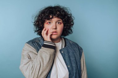 A young individual with curly hair stands in front of a light blue wall, showcasing a surprised expression while gently touching their face. They wear a casual jacket and a shirt.
