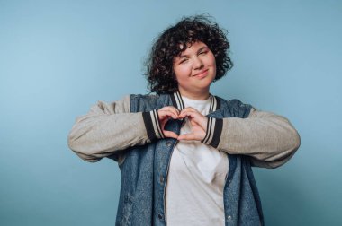 A young person smiles and forms a heart shape with their hands. They wear a relaxed outfit, showcasing a positive and friendly expression in a simple, pleasant environment.