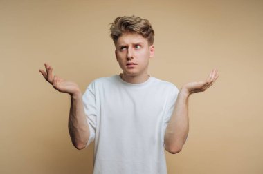 A young man stands in front of a smooth, light-colored wall, appearing puzzled as he gestures with his hands raised. His expression shows uncertainty or indecision.