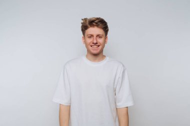 A young man stands smiling in a simple setting, wearing a white t-shirt. His cheerful demeanor adds warmth to the plain backdrop, creating an inviting atmosphere.