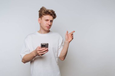 A young man stands in a bright, empty room, holding a smartphone and looking puzzled. He gestures with his other hand, indicating his confusion or curiosity about something.