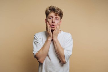 A young man stands with a surprised expression, hands on his face, against a light, neutral background. His casual white t-shirt suggests a relaxed indoor atmosphere.