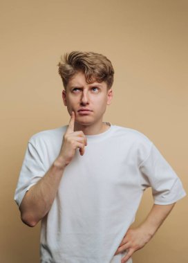 A young man stands with a pensive expression, finger on his chin, clearly lost in thought. He is dressed casually, and the background is a soft, muted color.