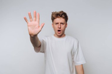 A young man with light hair stands against a plain background, raising his hand with a surprised expression. He seems to be signaling to stop or pause during a conversation.