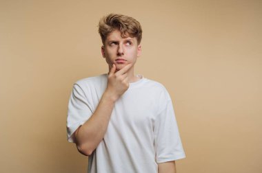 A young man stands with his hand on his chin, looking up with a thoughtful expression. He wears a plain white t-shirt and is in front of a soft beige backdrop, suggesting contemplation.