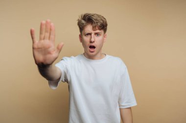 A young man stands with his palm raised, showing surprise or concern. His expression is intense, and the background is a soft, neutral color that emphasizes his reaction.