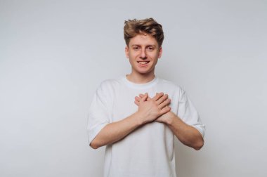 A young man stands in a studio against a plain, light backdrop. He smiles warmly while placing his hands on his chest, creating a friendly and inviting atmosphere.