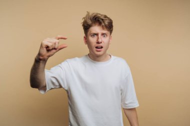 A young man gestures to indicate a small size with his fingers while expressing surprise or frustration. He wears a simple white shirt and stands against a neutral background.