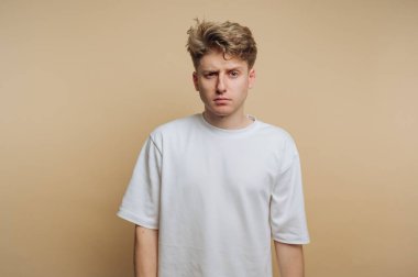 A young man stands with a neutral expression on his face, dressed in a plain white shirt. The background is a simple beige color, emphasizing his serious demeanor.