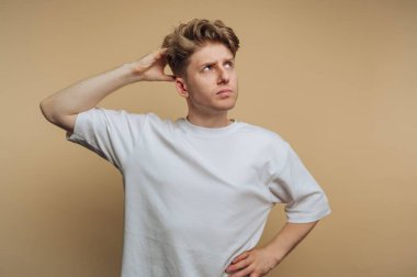 A young man stands with a curious look on his face, scratching his head thoughtfully. He wears a casual white shirt and seems to be pondering a question or dilemma.