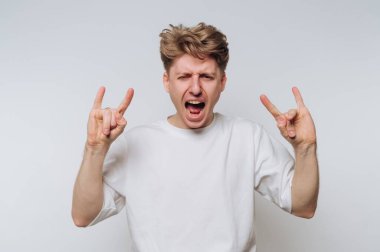 A young man with tousled hair shows his enthusiasm by raising his hands in a rock gesture. He is indoors against a plain background, conveying joy and liveliness.