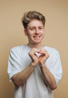 A cheerful young man forms a heart shape with his hands, displaying a friendly smile. He stands in front of a beige backdrop, creating a warm and inviting atmosphere.