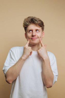 A young man with light hair shows a thoughtful expression while pointing at his cheeks. He is wearing a simple white shirt and stands against a neutral background.
