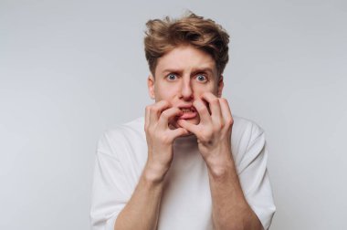 A young man with light brown hair displays a worried expression while gripping his face with both hands. The setting is simple, emphasizing his emotional state.