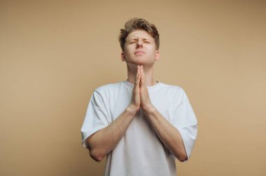 A young man stands with his eyes closed, hands pressed together in a prayer position. He appears to be reflecting or expressing gratitude. The simple background keeps the focus on his emotion.