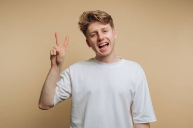 A young man with light brown hair smiles cheerfully while making a peace sign with his hand. He wears a simple white t-shirt and stands against a plain beige background.