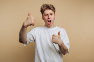 A young man stands in front of a plain light background, enthusiastically showing two thumbs up while making a joyful expression. His energy conveys optimism and excitement.