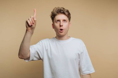 A young man wearing a plain white shirt gestures with his finger raised, expressing surprise or realization. He stands in front of a light beige background, looking upward.