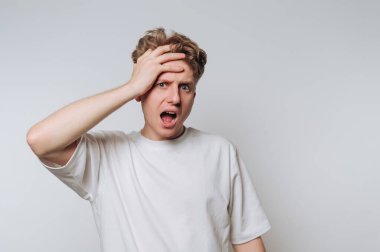 A young man stands against a plain white background, visibly shocked and frustrated. His hand rests on his forehead as he mouths an expression of disbelief at something unexpected.