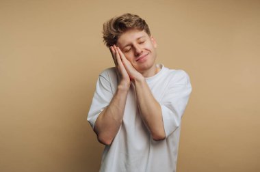 A young man stands against a warm beige background, with a smile on his face. He places his hands on his cheeks, showing feelings of happiness and tranquility.