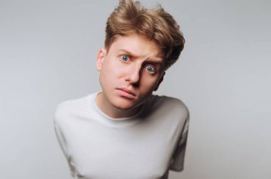 A young man with tousled light hair leans forward, showing a look of curiosity in a plain studio. The neutral background highlights his facial expression and bold blue eyes.