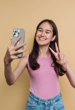 A young woman stands in front of a yellow wall, happily taking a selfie. She poses with a peace sign, wearing a pink top and denim shorts. Her smile conveys joy.