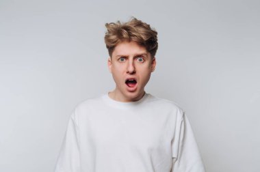 A young man with tousled hair shows a look of surprise and disbelief. He stands against a plain, light-colored background, dressed casually. His eyes are wide and mouth open.