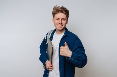 A young man stands in a light-colored room, smiling and giving a thumbs up. He holds a laptop close to his body, wearing a casual outfit.