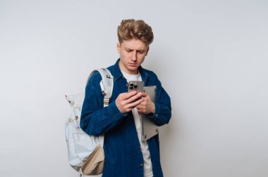 A young student stands indoors, focused on his smartphone while holding a laptop under his arm. He wears casual clothing and a backpack, appearing engaged in his device.