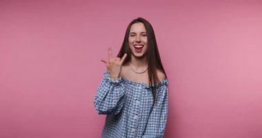 footage of young woman with long hair dressed in a blue checked dress, playfully making rock signs while showing a fun expression on pink background