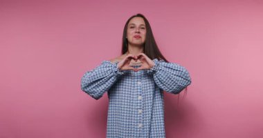 footage of young woman wearing a blue dress smiling widely while making a heart gesture with her fingers, exuding happiness on pink background