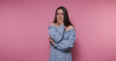 footage of young woman in a checkered blue dress poses, expressing emotion while embracing herself on pink background