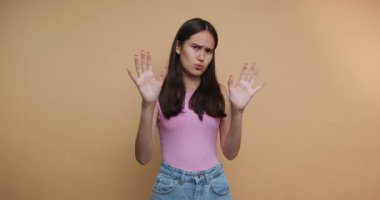 footage of young woman stands against a pastel background, raising her hands in refusal, capturing a moment of playful defiance on beige background